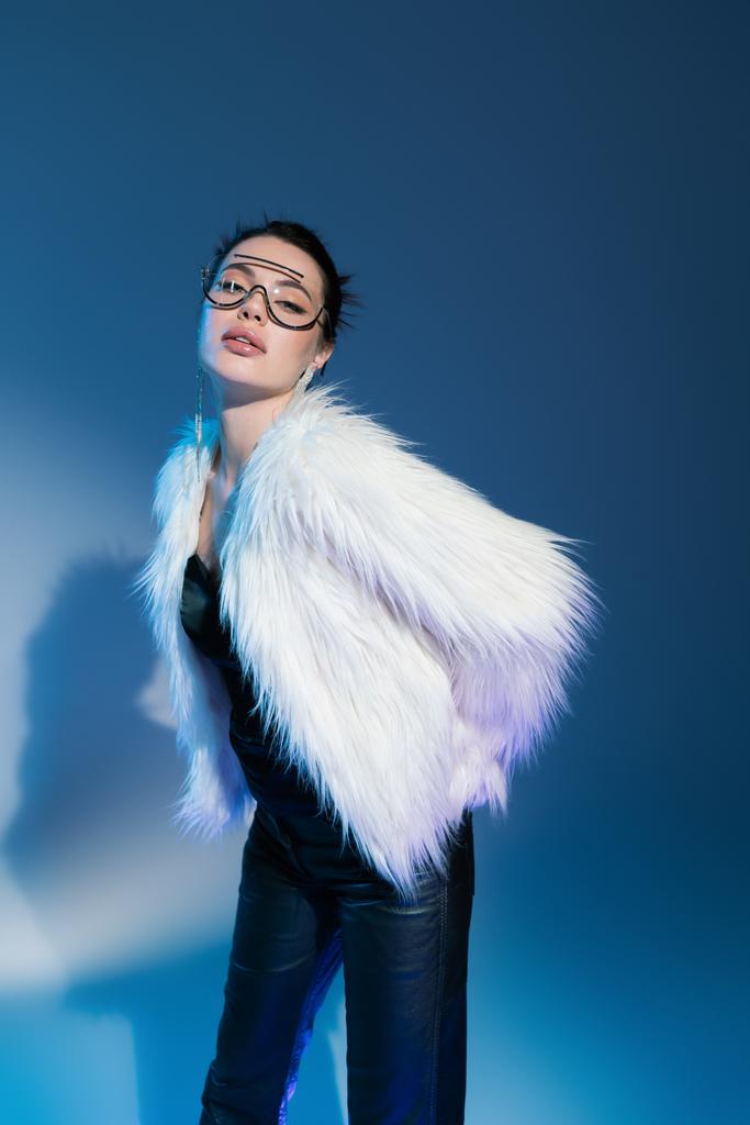 young brunette woman in white faux fur jacket and transparent sunglasses looking at camera while posing on blue background - Photo, Image