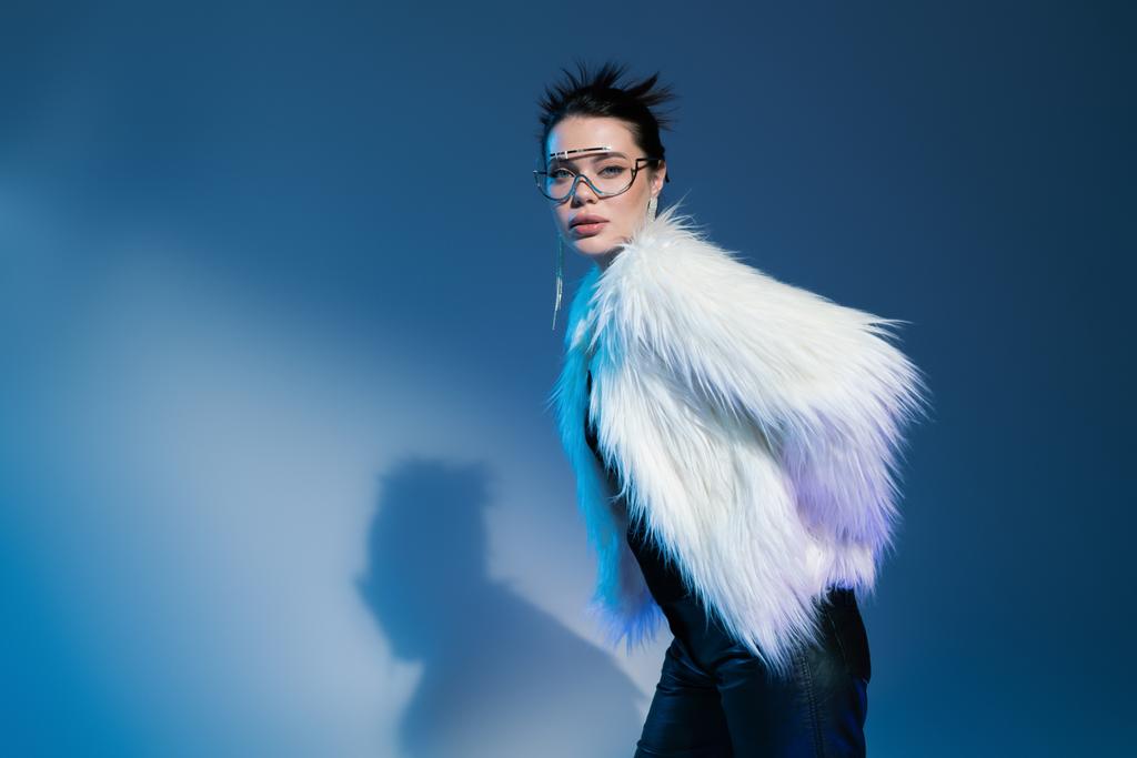 young woman in transparent eyeglasses and fluffy faux fur jacket looking at camera on blue background - Photo, Image