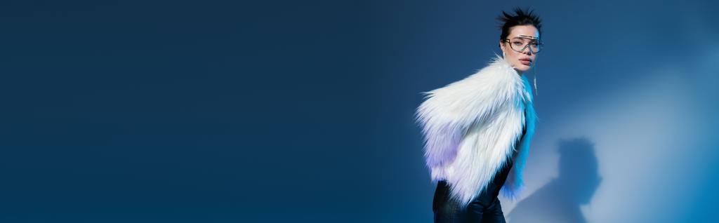 modna kobieta w stylowej, sztucznej kurtce i przezroczystych okularach przeciwsłonecznych na niebieskim tle z przestrzenią do kopiowania, baner - Zdjęcie, obraz