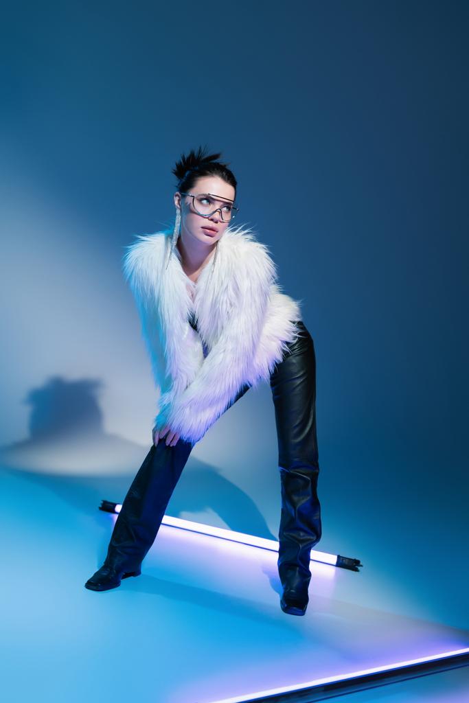 modelo morena en pantalones de cuero negro y chaqueta de piel sintética blanca mirando hacia otro lado cerca de la lámpara fluorescente sobre fondo azul - Foto, Imagen
