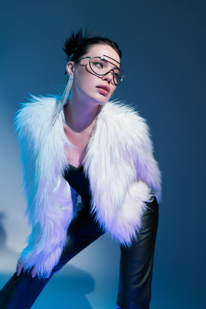 young model in transparent eyeglasses and white faux fur jacket looking away on blue background - Photo, Image