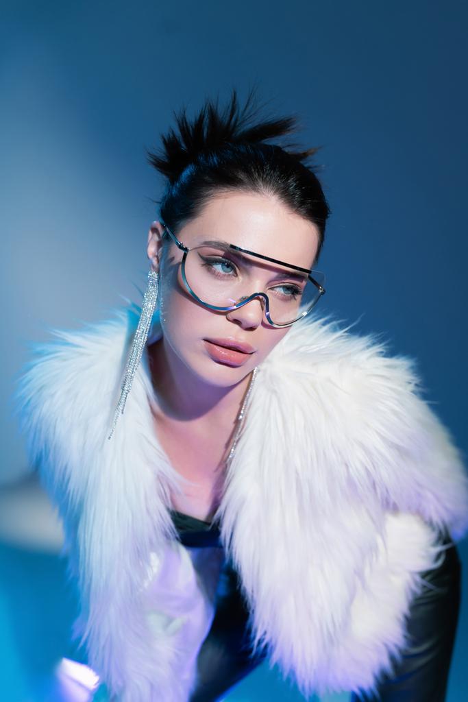portrait of charming woman in white faux fur jacket and transparent sunglasses looking away on blue background - Photo, Image
