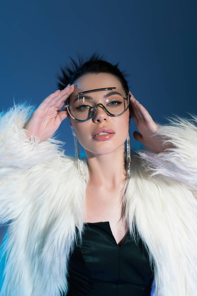 divatos nő bolyhos műszőrme kabát és átlátszó napszemüveg fogja kezét közel arc kék háttér - Fotó, kép