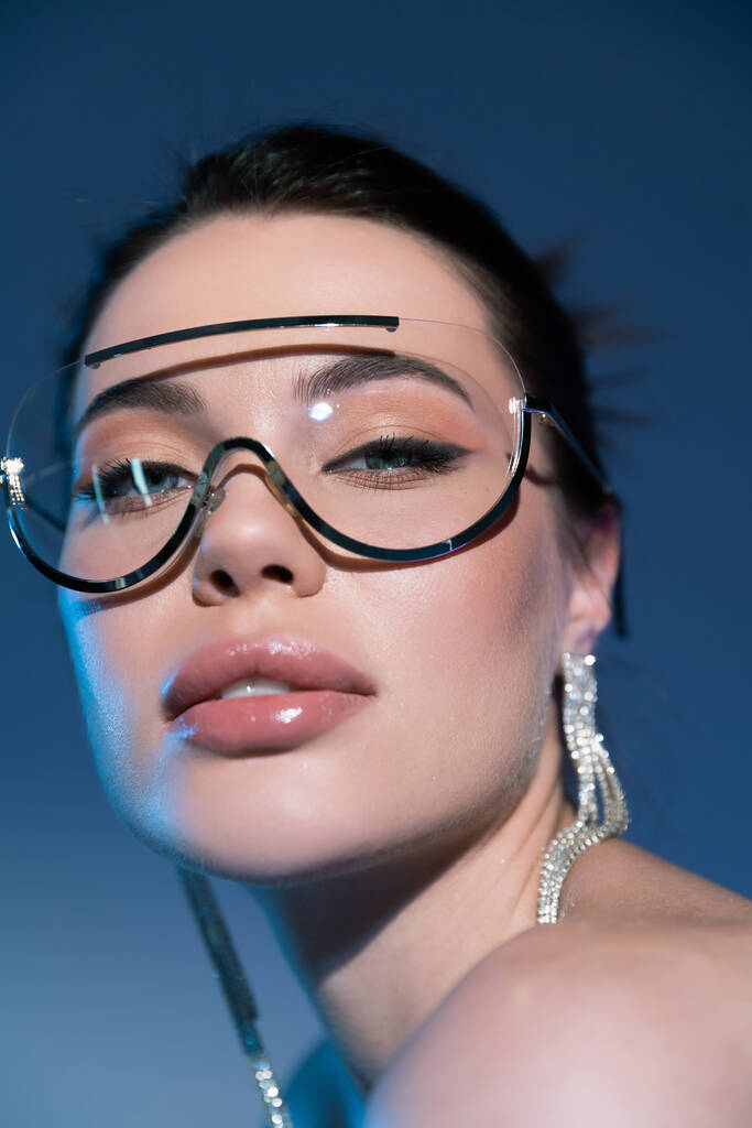 zblízka portrét ženy v make-upu a stylové sluneční brýle při pohledu na kameru na modrém pozadí - Fotografie, Obrázek