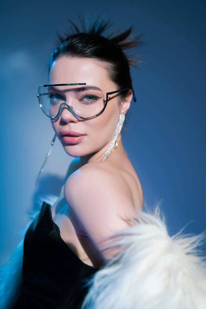 trendy woman in black corset and transparent sunglasses posing with faux fur jacket on blue background - Photo, Image