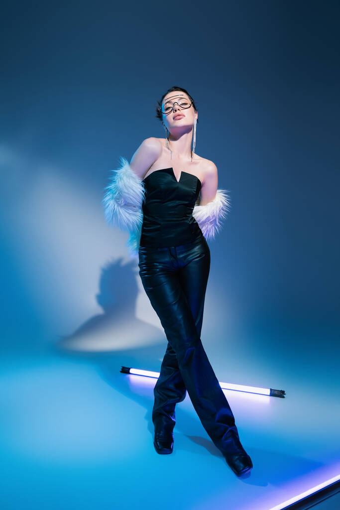 volledige lengte van trendy vrouw in zwart lederen broek en witte harige jas staande met gekruiste benen in de buurt lamp op blauwe achtergrond - Foto, afbeelding