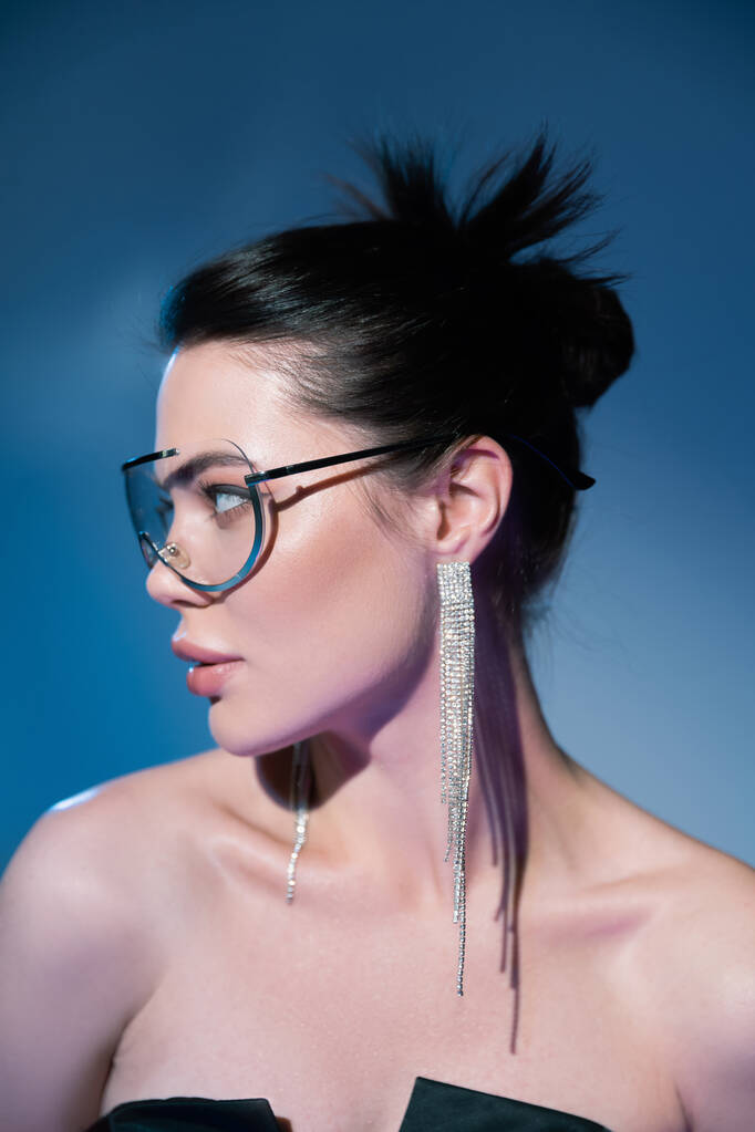 πορτρέτο της μελαχρινής γυναίκας στη μόδα διαφανή γυαλιά κοιτάζοντας μακριά στο μπλε φόντο - Φωτογραφία, εικόνα