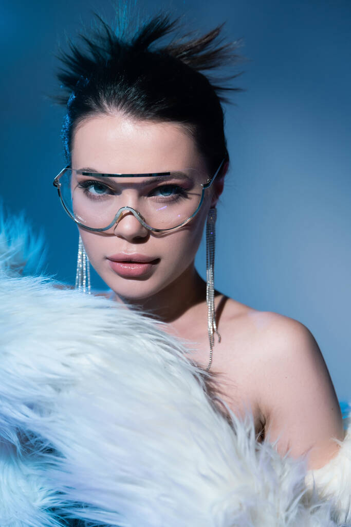 retrato de modelo glamour en gafas de sol transparentes y pendientes brillantes posando cerca de chaqueta de piel sintética blanca sobre fondo azul - Foto, Imagen