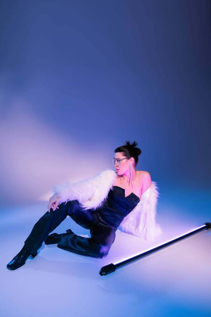 glamour woman in black corset and white furry jacket sitting near fluorescent lamp on blue background - Photo, Image