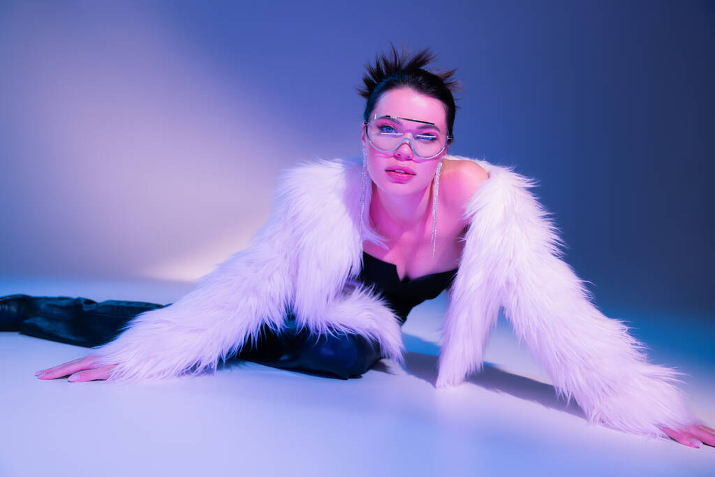 seductive woman in white faux fur jacket and transparent sunglasses posing on blue and purple background - Photo, Image
