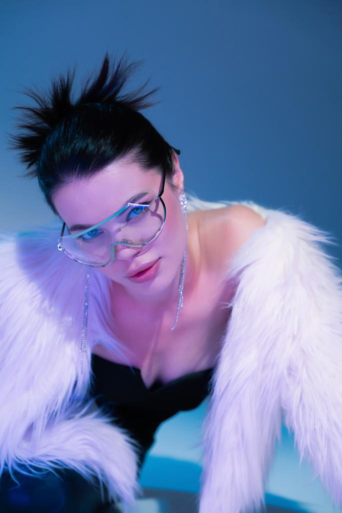 fashionable woman in white faux fur jacket and transparent eyeglasses on blue background with purple light - Photo, Image