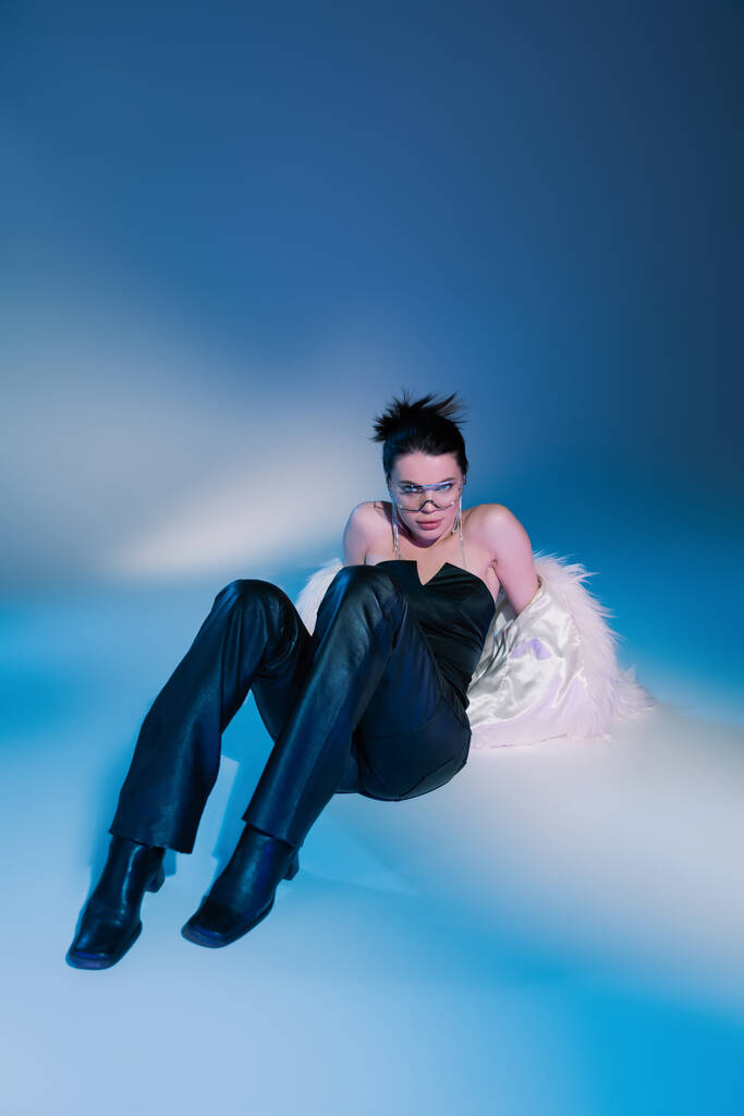full length of stylish woman in white faux fur jacket and black leather pants posing on blue background - Photo, Image