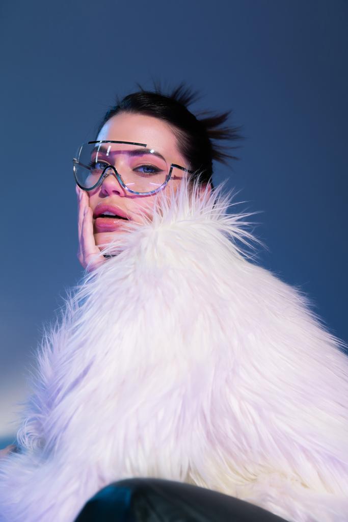 αισθησιακό μελαχρινό μοντέλο σε λευκό faux γούνα σακάκι και διάφανα γυαλιά ηλίου αγγίζοντας το πρόσωπο σε μπλε φόντο - Φωτογραφία, εικόνα