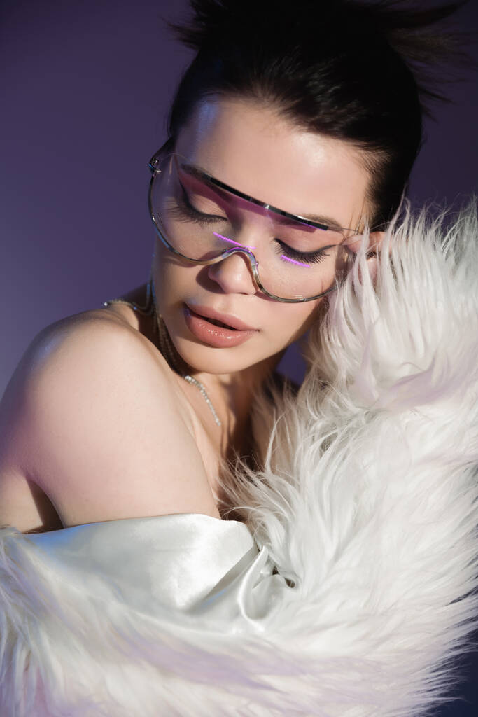 érzéki barna nő divatos szemüveg és fehér műszőrme kabát pózol lila háttér - Fotó, kép