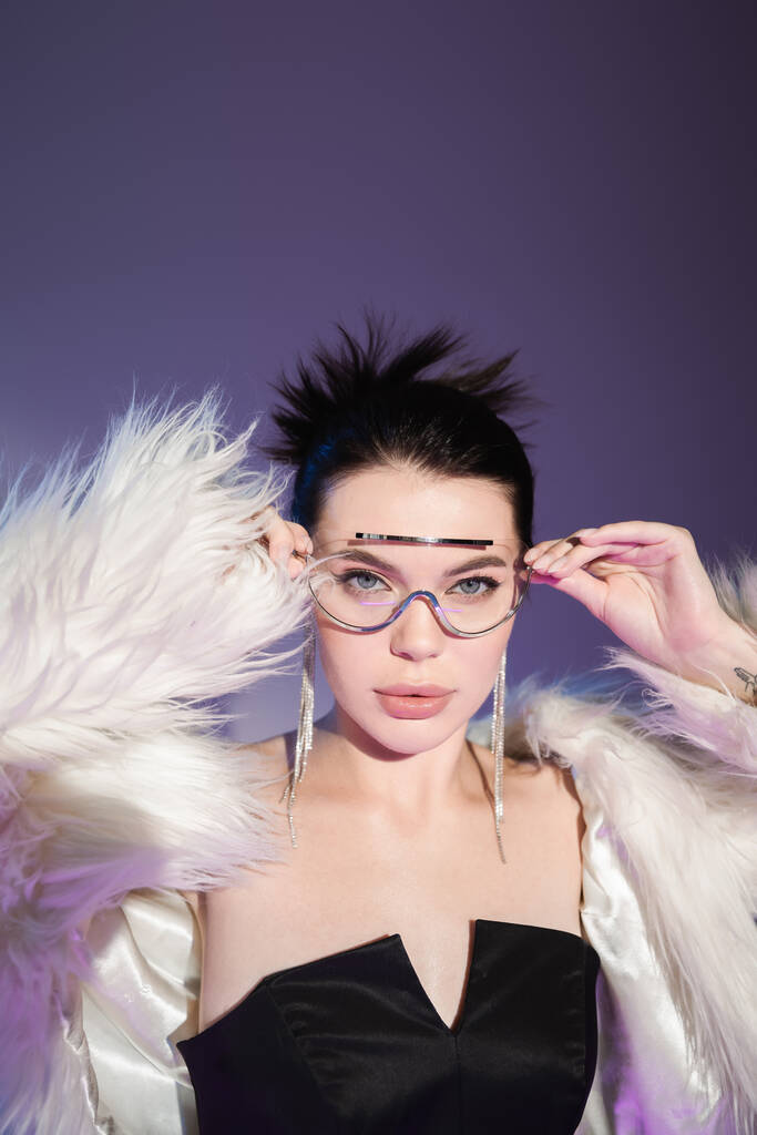 modelo glamour en chaqueta de piel sintética blanca ajustando gafas de sol transparentes sobre fondo púrpura - Foto, Imagen