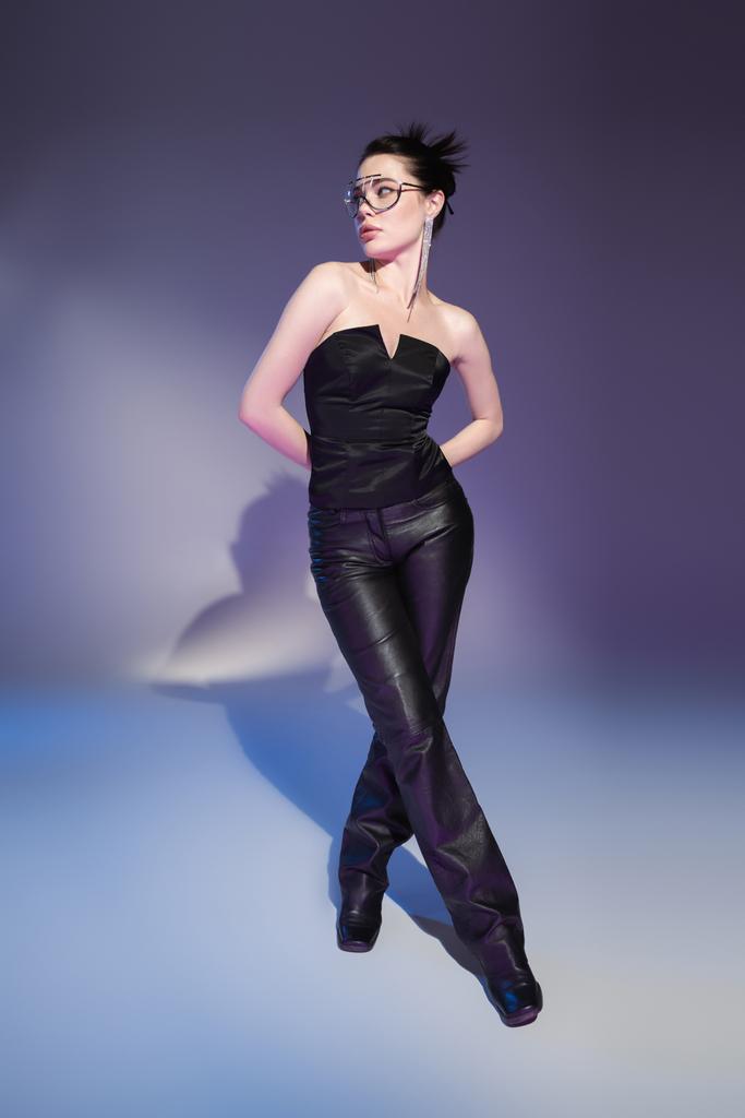 Полная длина гламурная женщина в кожаных штанах и черный корсет позирует с руками сзади на фиолетовом фоне - Фото, изображение