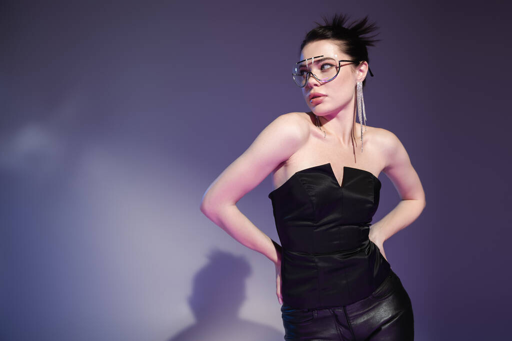 seductive woman in black corset and trendy sunglasses standing with hands on hips on purple background - Photo, Image