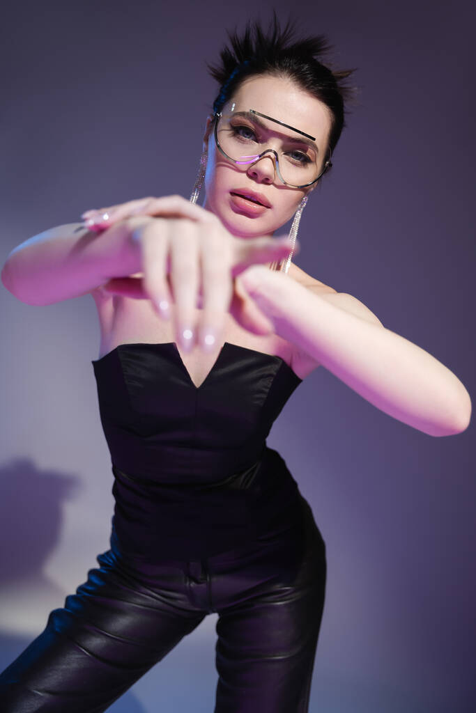 brünette Frau in schwarzem Korsett und trendiger Sonnenbrille blickt in die Kamera und zeigt mit dem Finger auf lila Hintergrund - Foto, Bild