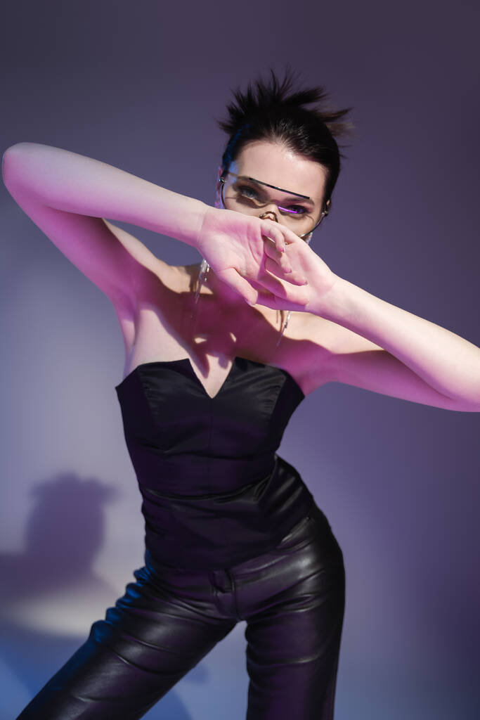 brunette woman in black corset and leather trousers obscuring face with hands on purple background - Photo, Image