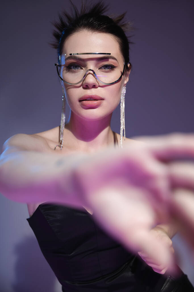 гламурная женщина в прозрачных солнцезащитных очках, смотрящая на камеру рядом с размытой протянутой рукой на фиолетовом фоне - Фото, изображение