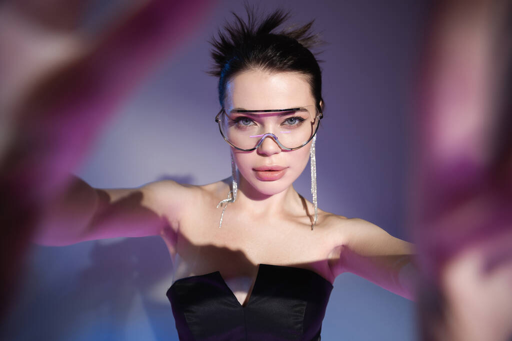 Glamour-Frau mit transparenter Brille und schwarzem Korsett blickt in die Kamera auf verschwommenem lila Hintergrund - Foto, Bild