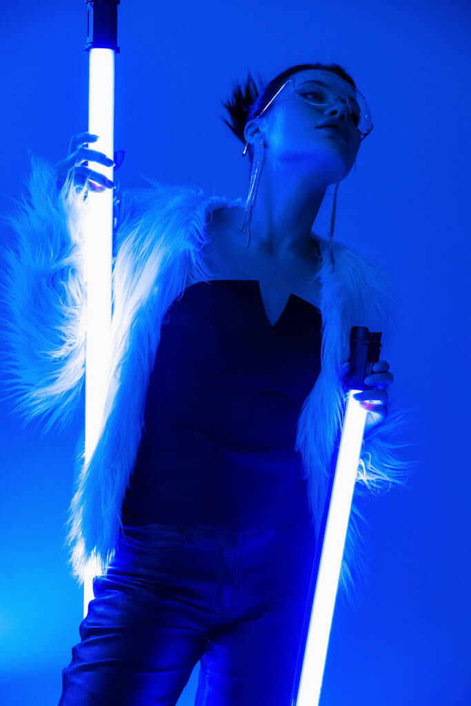 low angle view of trendy woman in faux fur jacket and black corset holding fluorescent lamps on blue background - Photo, Image