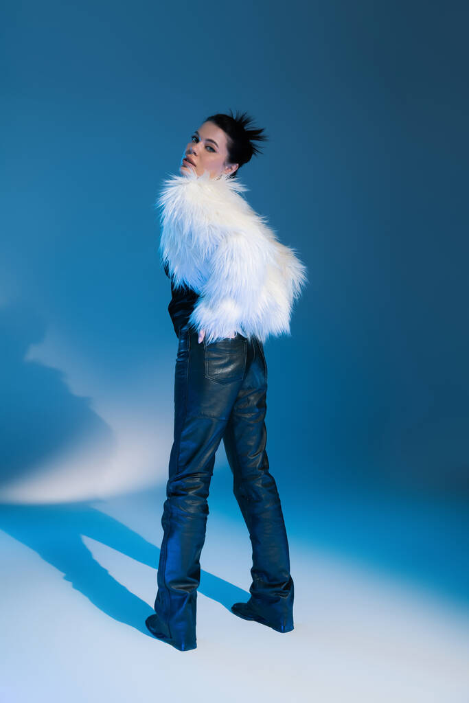 Full length of trendy model in faux fur jacket looking at camera on blue background  - Photo, Image