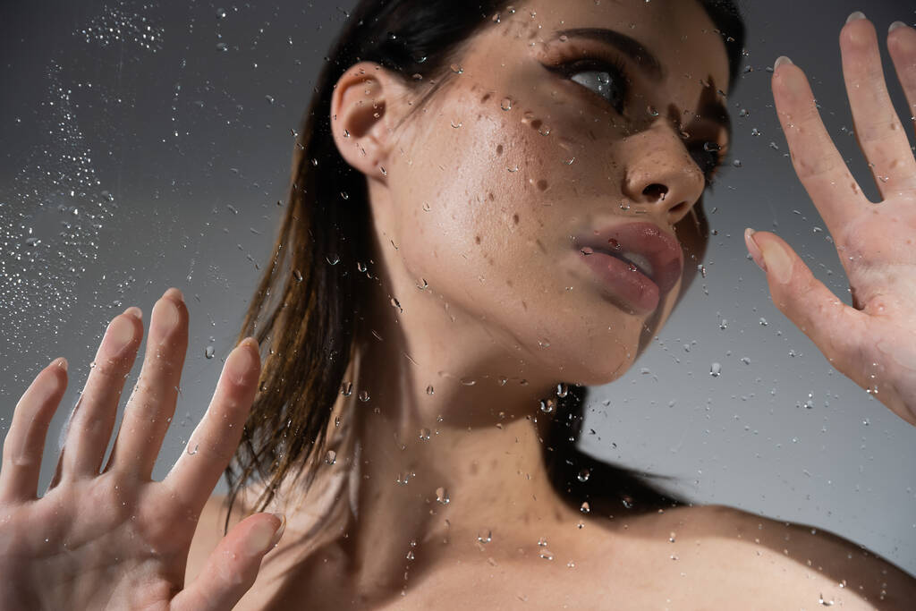 Low angle view of young brunette model with naked shoulders touching wet glass on grey background  - Photo, Image