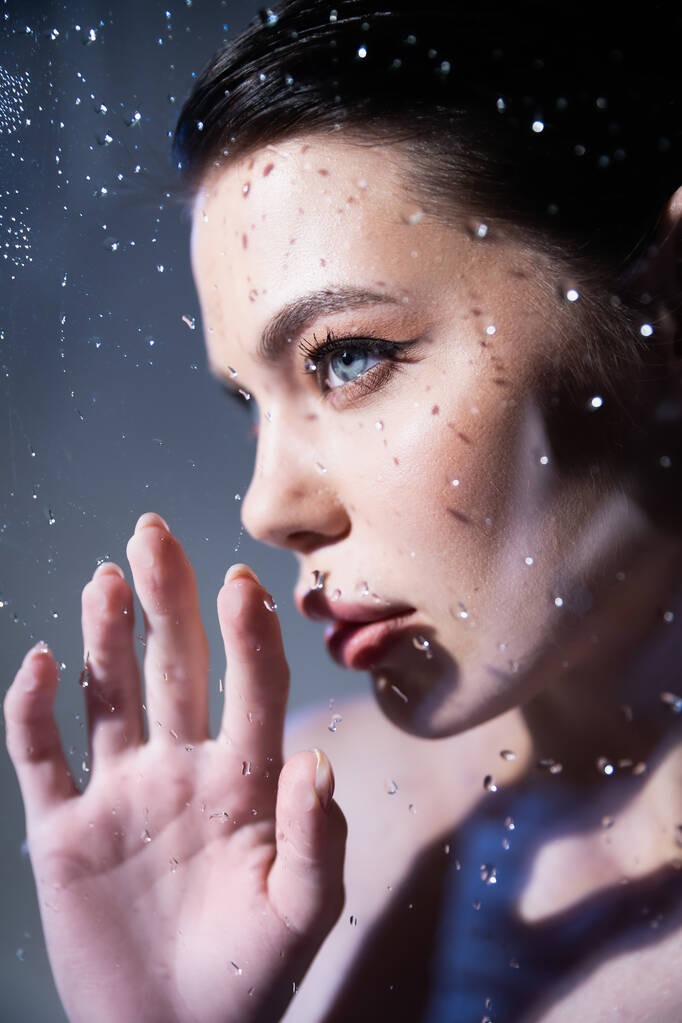 Young model with makeup touching glass with water drops on grey background  - Photo, Image
