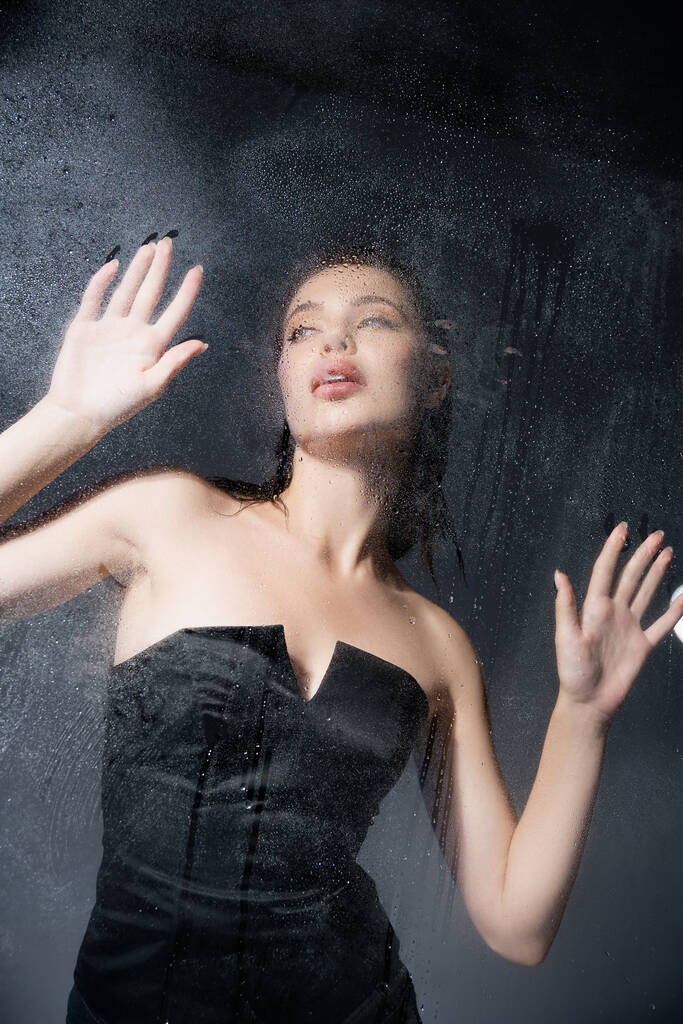 Low angle view of fashionable woman in corset standing behind wet glass on grey background  - Photo, Image