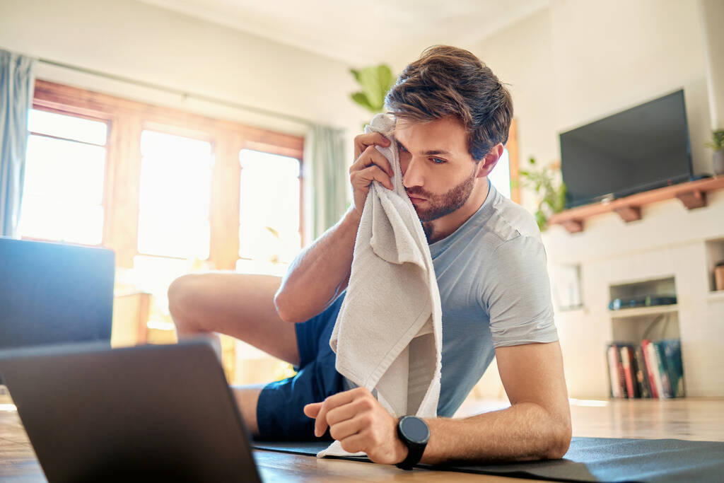Ein fitter junger Mann wischt sich mit einem Handtuch den Schweiß von der Stirn, während er zu Hause eine Trainingspause einlegt. Kerl sieht müde aus beim Training mit Online-Workout-Tutorials auf digitalem Tablet. - Foto, Bild