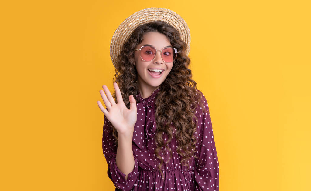 glückliches Kind mit Strohhut und Sonnenbrille mit langen brünetten lockigen Haaren auf gelbem Hintergrund. - Foto, Bild