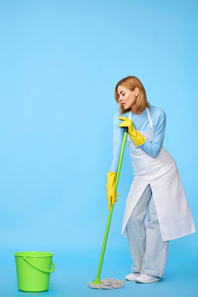 Lastik eldivenli ve temiz önlüklü mutlu bir kadın mavi arka planda paspasla yerleri yıkıyor. Tam uzunluk - Fotoğraf, Görsel