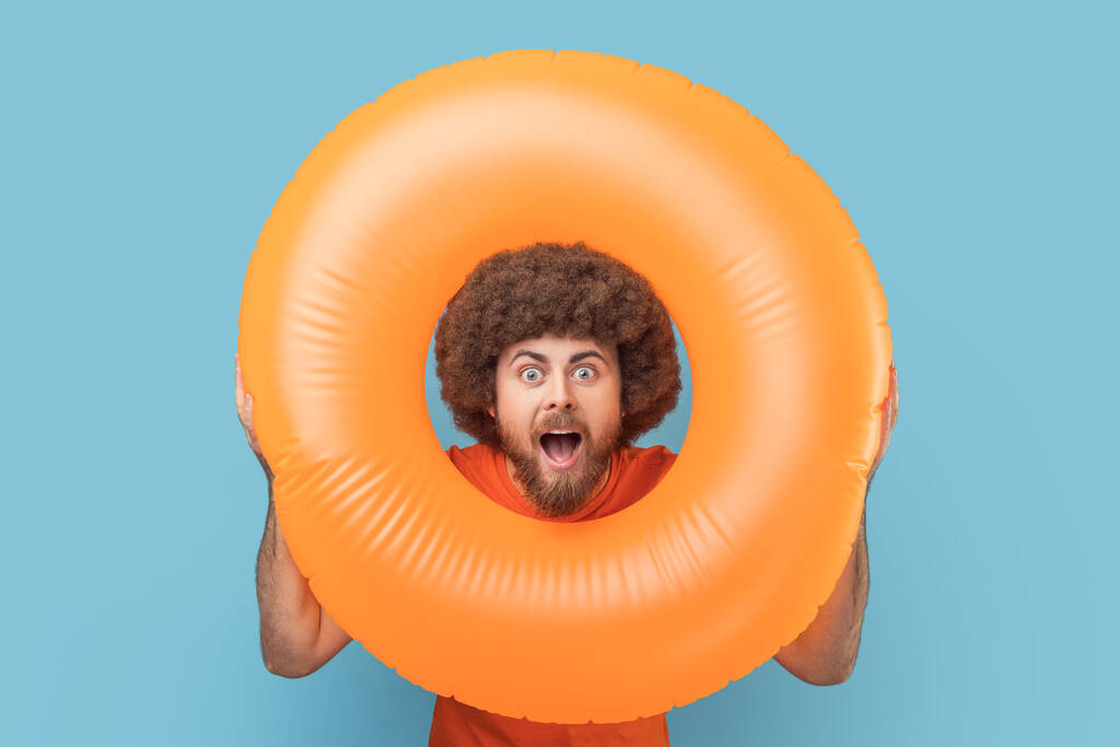 Portrait of man with shocked facial expression looking trough bright orange rubber ring, looking at camera with open mouth, surprised by vacation. Indoor studio shot isolated on blue background. - Photo, Image