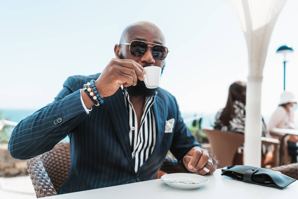 Güneş gözlüklü şık kel bir Afrikalı adamın portresi ve şık bir yaz elbisesi bir kafede oturuyor ve kahve içiyor, elinde bir fincan espresso ile seçici bir odak noktası var. - Fotoğraf, Görsel