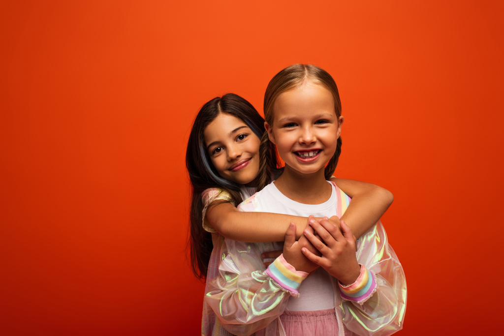 brunetka dítě objímající šťastný přítel a usmívat se na kameru izolované na oranžové - Fotografie, Obrázek