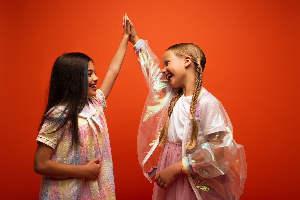 niñas felices en el vestido y chaqueta de lluvia transparente dando alta cinco aislado en naranja - Foto, Imagen