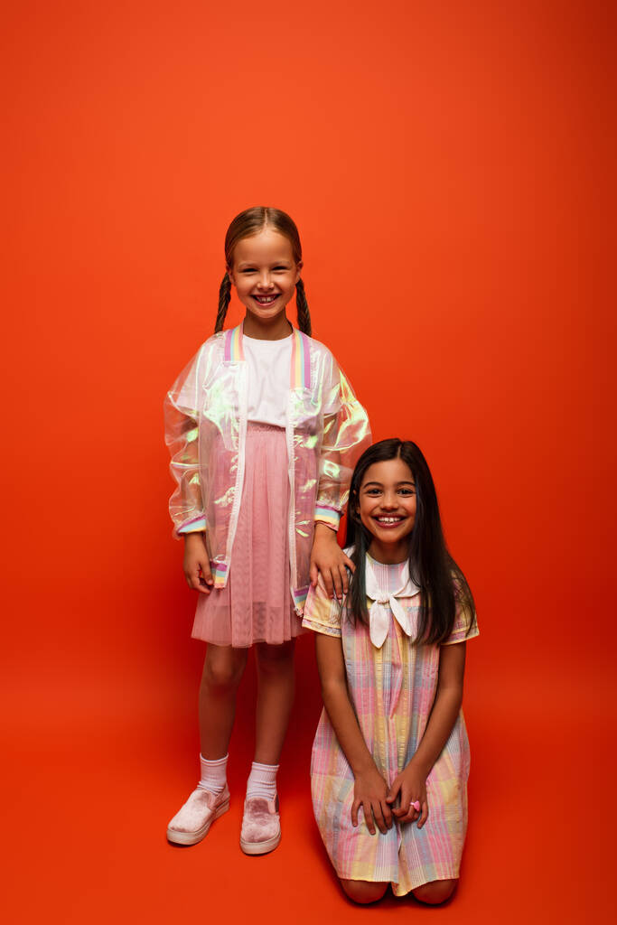 brünettes Mädchen im Kleid sitzt neben lächelnden Freund in Regenjacke auf orangefarbenem Hintergrund - Foto, Bild