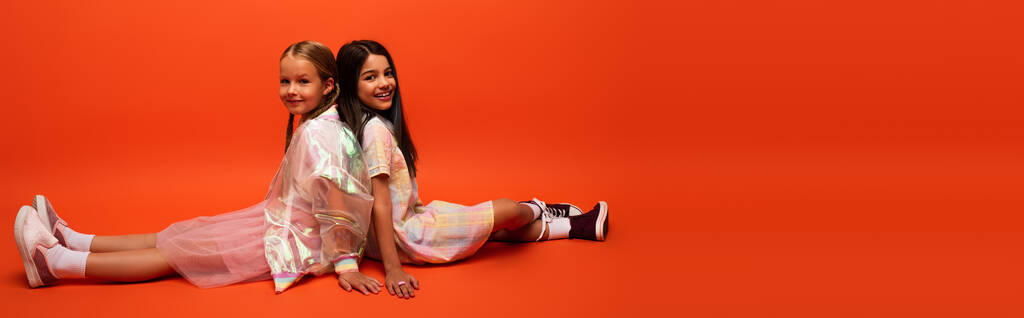 full length of happy and trendy girls sitting back to back and looking at camera on orange background, banner - Photo, Image