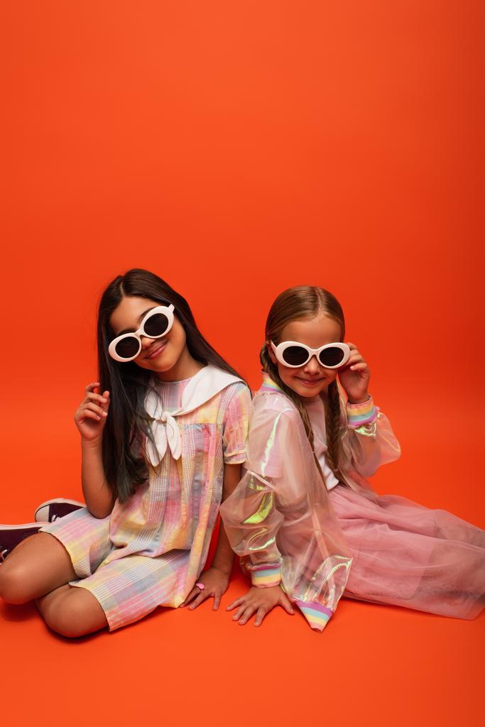 mosolygós tizenéves lányok divatos ruhák és napszemüveg nézi a kamera, miközben ül a narancssárga háttér - Fotó, kép