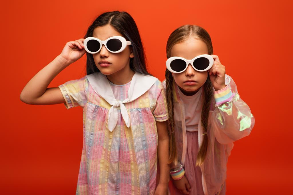 dívky s vážnými výrazy obličeje upravující sluneční brýle izolované na oranžové - Fotografie, Obrázek
