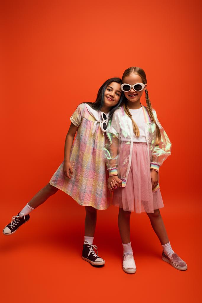 volle Länge des glücklichen Mädchens mit Sonnenbrille hält Händchen mit Freund, während sie auf orangefarbenem Hintergrund posiert - Foto, Bild