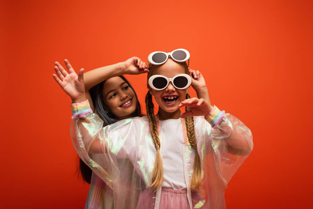vzrušená dívka ve dvou slunečních brýlích mává rukou a zároveň se baví s přítelem izolované na oranžové - Fotografie, Obrázek