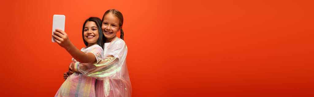 брюнетка делает селфи с другом в дождевой куртке обнимая ее изолированы на оранжевый, баннер - Фото, изображение