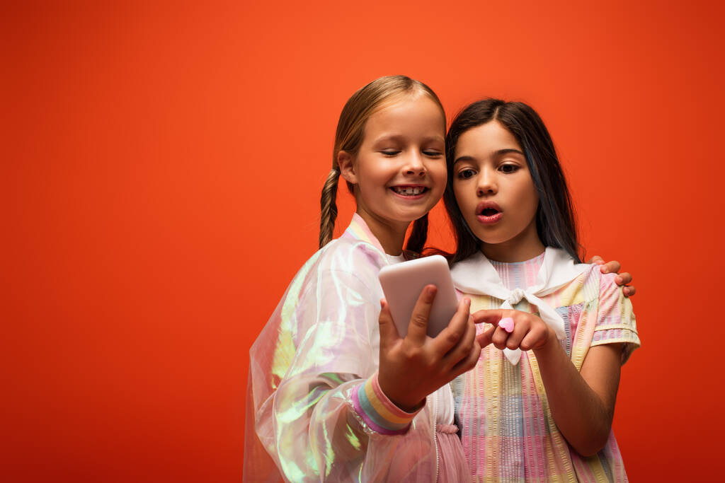 verbaasd meisje wijzen naar mobiele telefoon in de buurt glimlachende vriend geïsoleerd op oranje - Foto, afbeelding