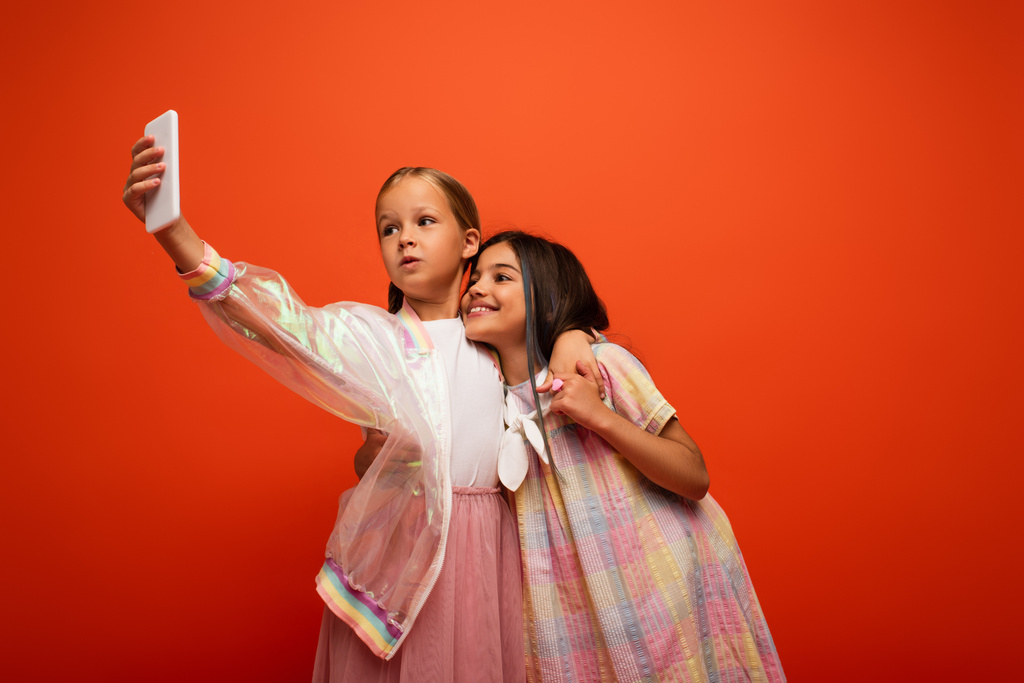Mädchen in transparenter Regenjacke macht Selfie mit glücklicher brünetter Freundin isoliert auf orange - Foto, Bild