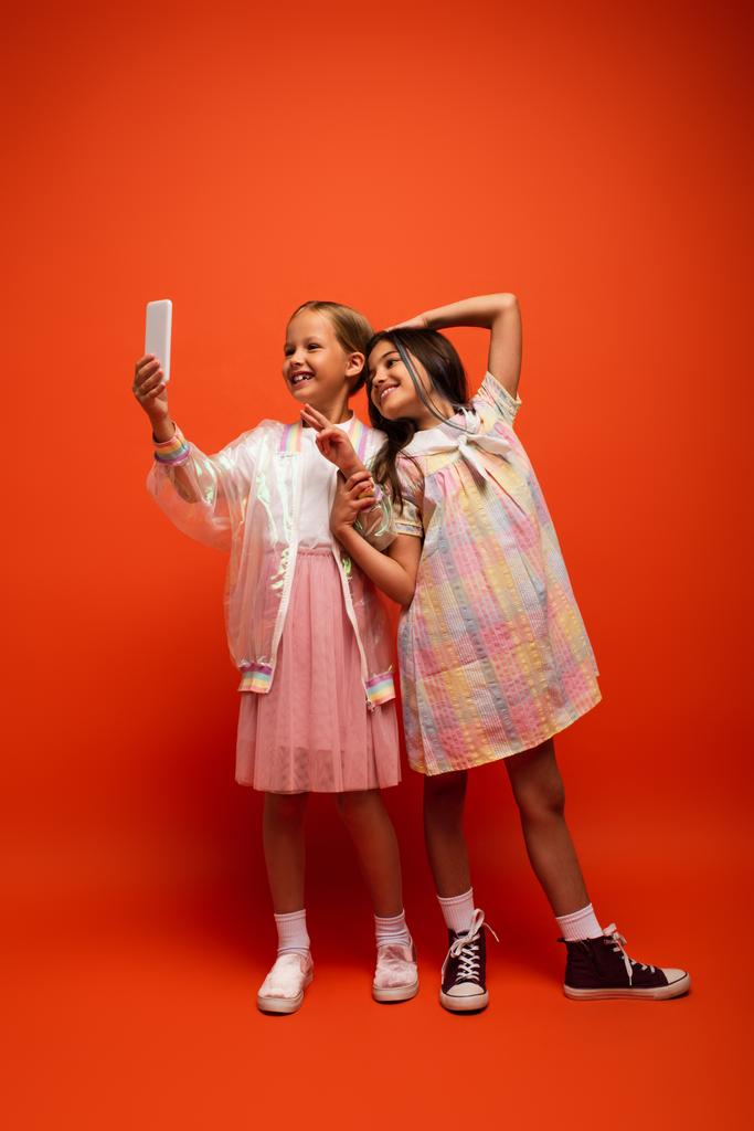 glückliches Mädchen zeigt Siegeszeichen in der Nähe Freund macht Selfie auf Handy auf orangefarbenem Hintergrund - Foto, Bild