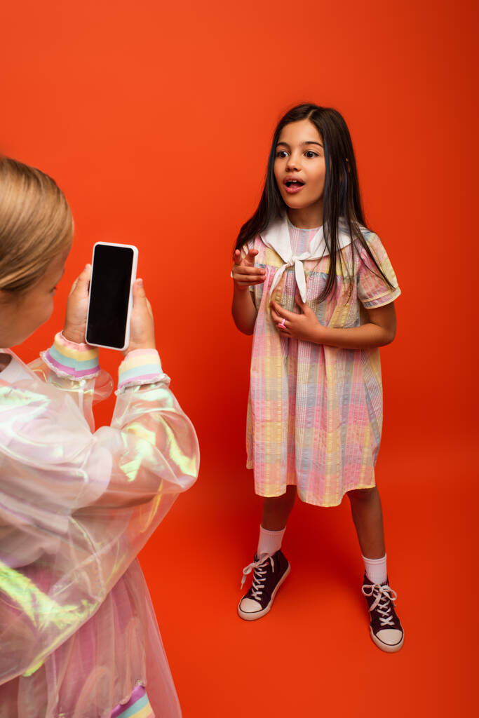 užaslá dívka mluví a ukazuje prstem v blízkosti přítele nahrávání videa na smartphone na oranžovém pozadí - Fotografie, Obrázek