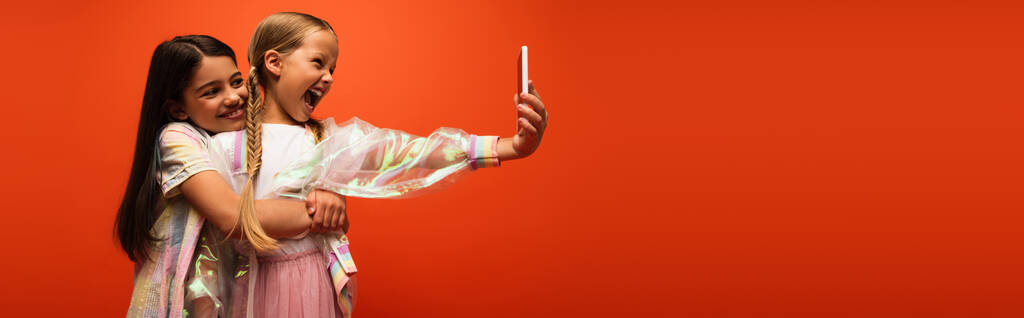 uśmiechnięta dziewczyna obejmująca podekscytowaną przyjaciółkę z otwartymi ustami robiącą selfie na smartfonie odizolowanym na pomarańczowo, baner - Zdjęcie, obraz