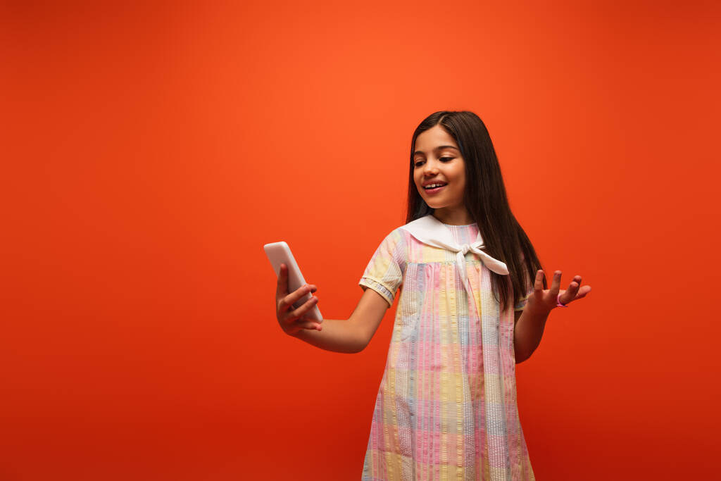 veselé brunetky dívka v šatech přičemž selfie na mobilu a ukazuje s rukou izolované na oranžové - Fotografie, Obrázek